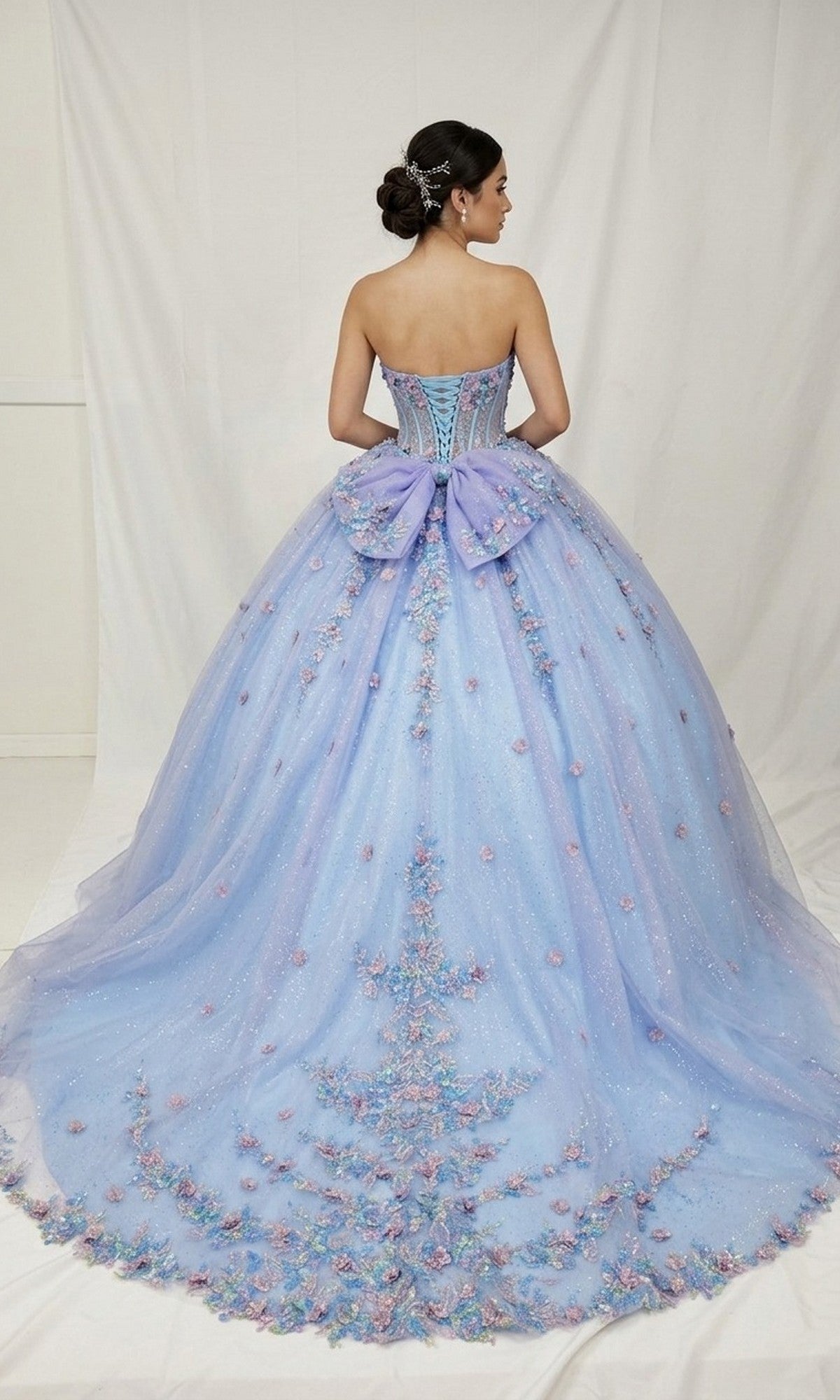 A woman wearing the Quinceanera Dress CHF3114 By Chicas—a strapless lavender ball gown accented with 3D floral appliques and a large bow—stands with her back to the camera against a plain white backdrop.