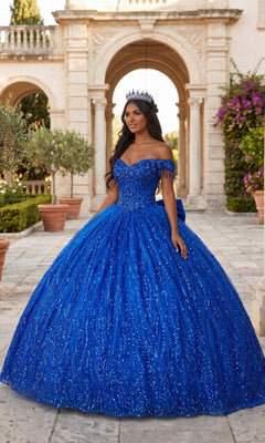 A woman stands outdoors in front of a grand, classical building with arches and lush greenery, wearing the Quinceanera Dress CHF3116 By Chicas in royal. The sparkling gown features a corset bodice and a flowing ballgown skirt, and she completes the enchanting scene with a crown.