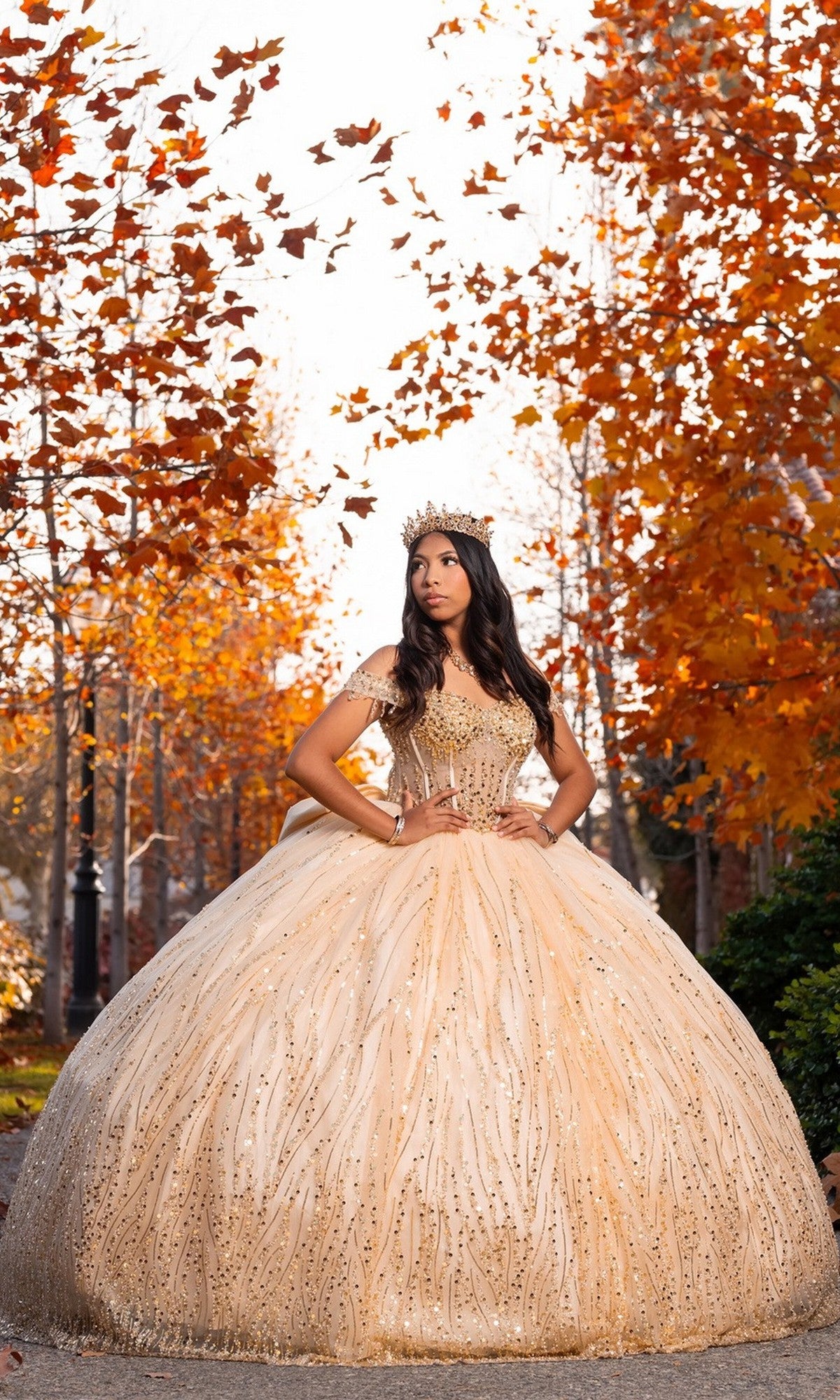 A young woman stands outdoors amid autumn trees, orange leaves gently falling around her, wearing the Quinceanera Dress CHF3116 By Chicas. The sparkling gown features a corset bodice and an elegant ballgown skirt, while a delicate crown graces her head, completing the enchanting scene.