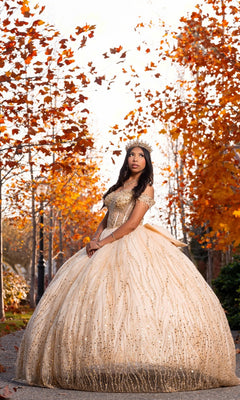 A young woman wearing the Quinceanera Dress CHF3116 By Chicas—a lavish gold ballgown skirt—paired with a tiara, stands outdoors amid autumn trees as orange leaves fall around her.