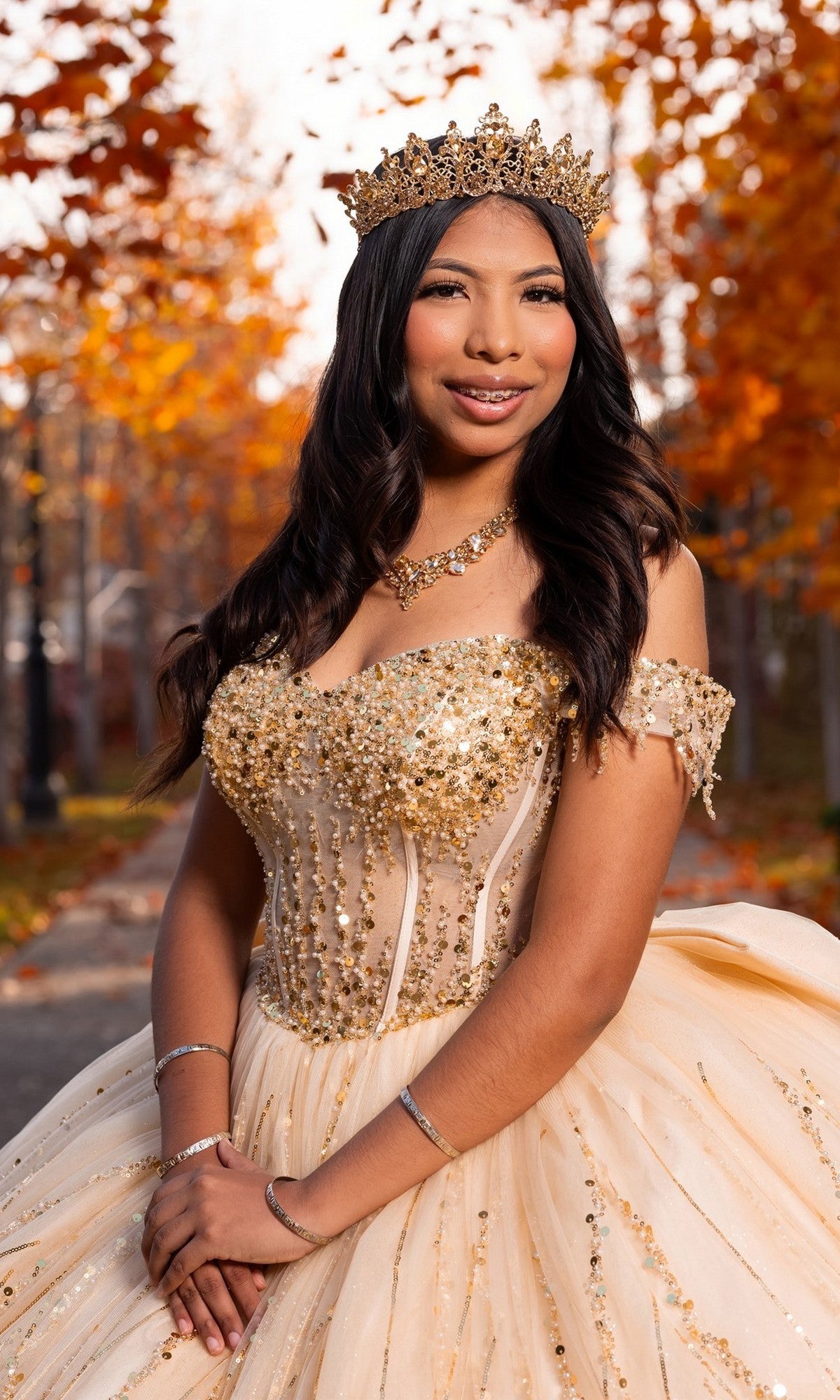 A young woman wearing the Quinceanera Dress CHF3116 By Chicas—a gold jeweled gown with a corset bodice and crown—stands outdoors on a path lined with autumn trees, smiling at the camera.