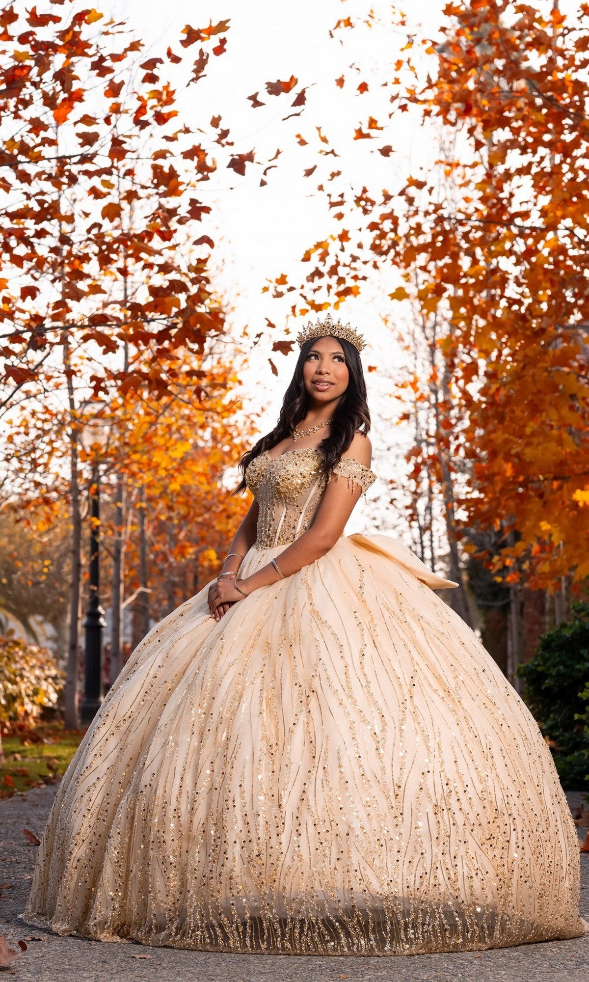 A young woman stands outdoors on a path framed by autumn trees and orange leaves, wearing the Quinceanera Dress CHF3116 By Chicas—a gold ball gown with a corset bodice and tiara.