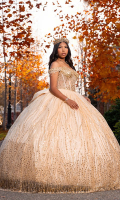 A young woman in the Quinceanera Dress CHF3116 By Chicas, featuring a sparkling champagne hue and corset bodice, stands outdoors wearing a tiara, framed by autumn trees and swirling orange leaves in the background.