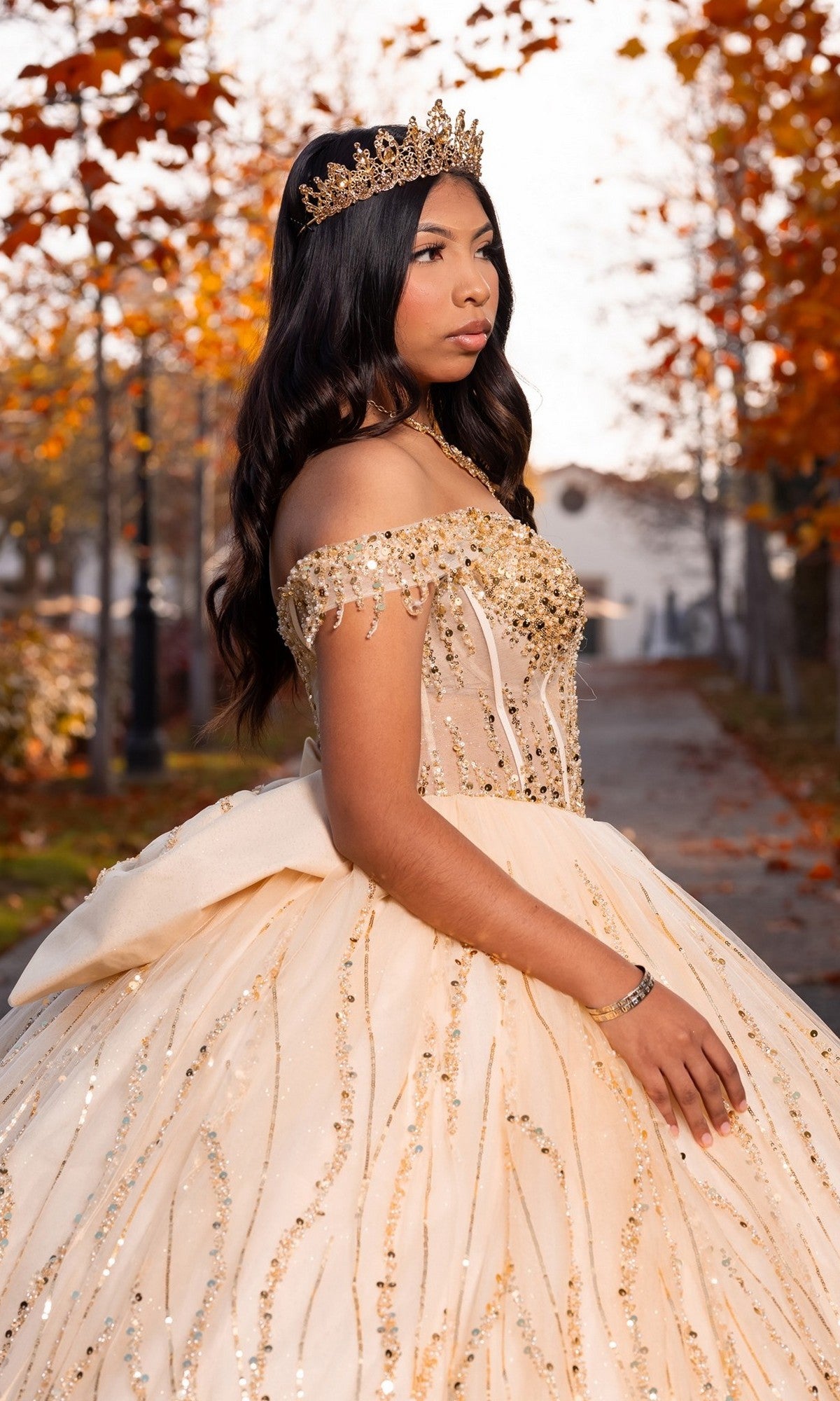 A young woman stands outdoors on a path lined with autumn trees, wearing the Quinceanera Dress CHF3116 By Chicas—an elegant beaded off-shoulder gown with a corset bodice—and a crown.
