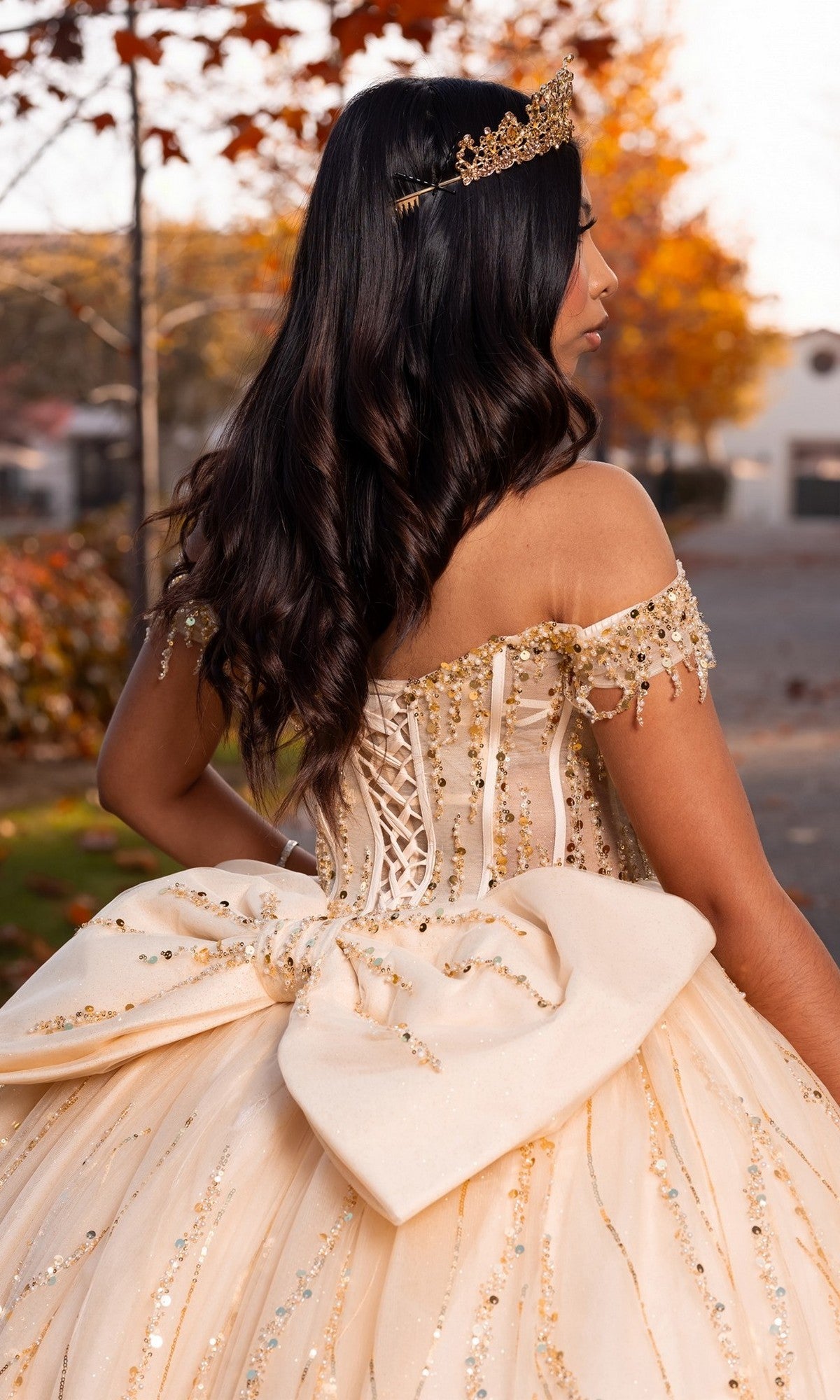 A woman is outdoors wearing the Quinceanera Dress CHF3116 By Chicas, an ornate off-the-shoulder cream gown embellished with gold sequins. The dramatic ballgown skirt, oversized back bow, and a sparkling gold tiara complete her look. Autumn leaves and softly blurred houses fill the background.