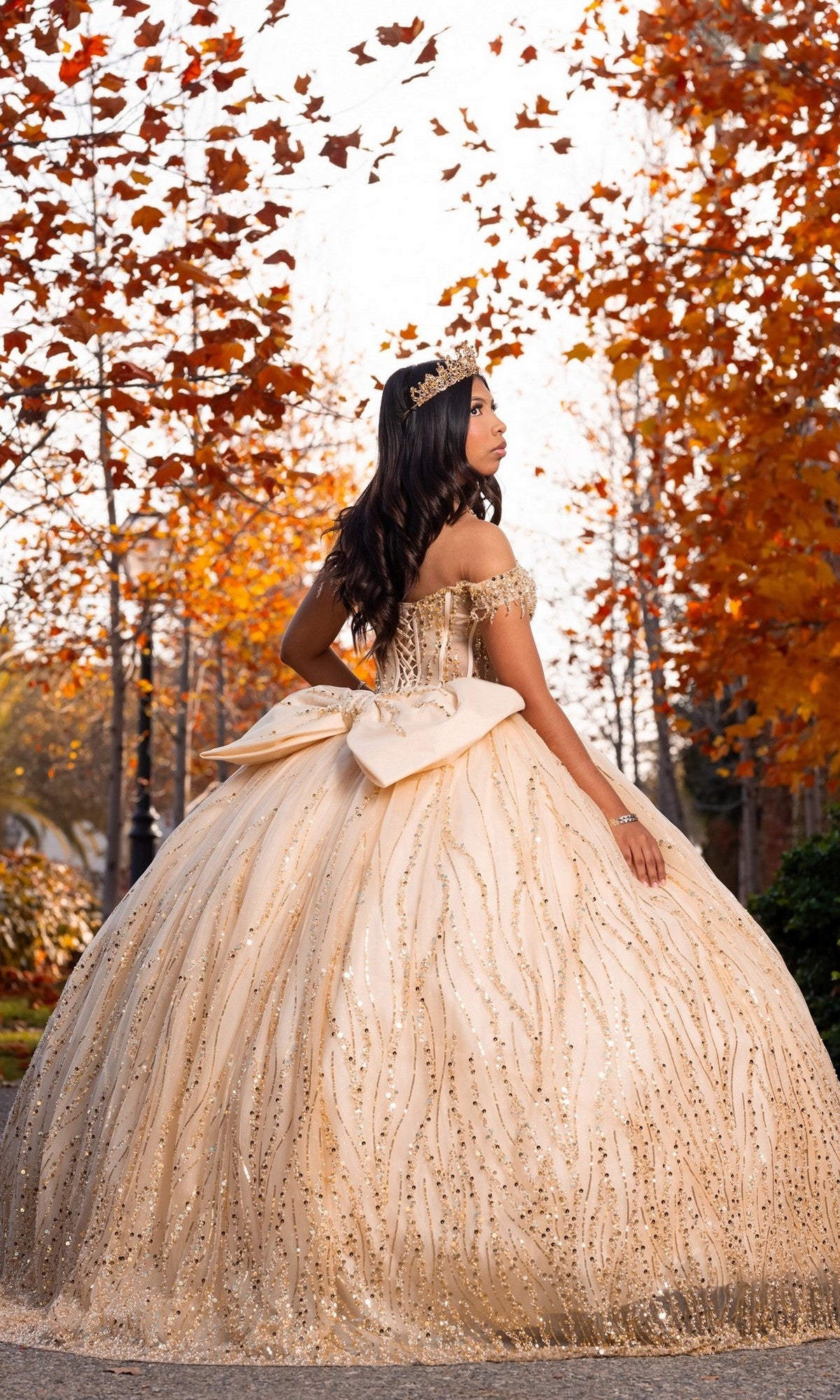 A young woman wearing the Quinceanera Dress CHF3116 By Chicas, featuring a gold off-shoulder corset bodice and dramatic ballgown skirt, stands outdoors among autumn trees with orange leaves, her tiara sparkling softly in the gentle fall light.