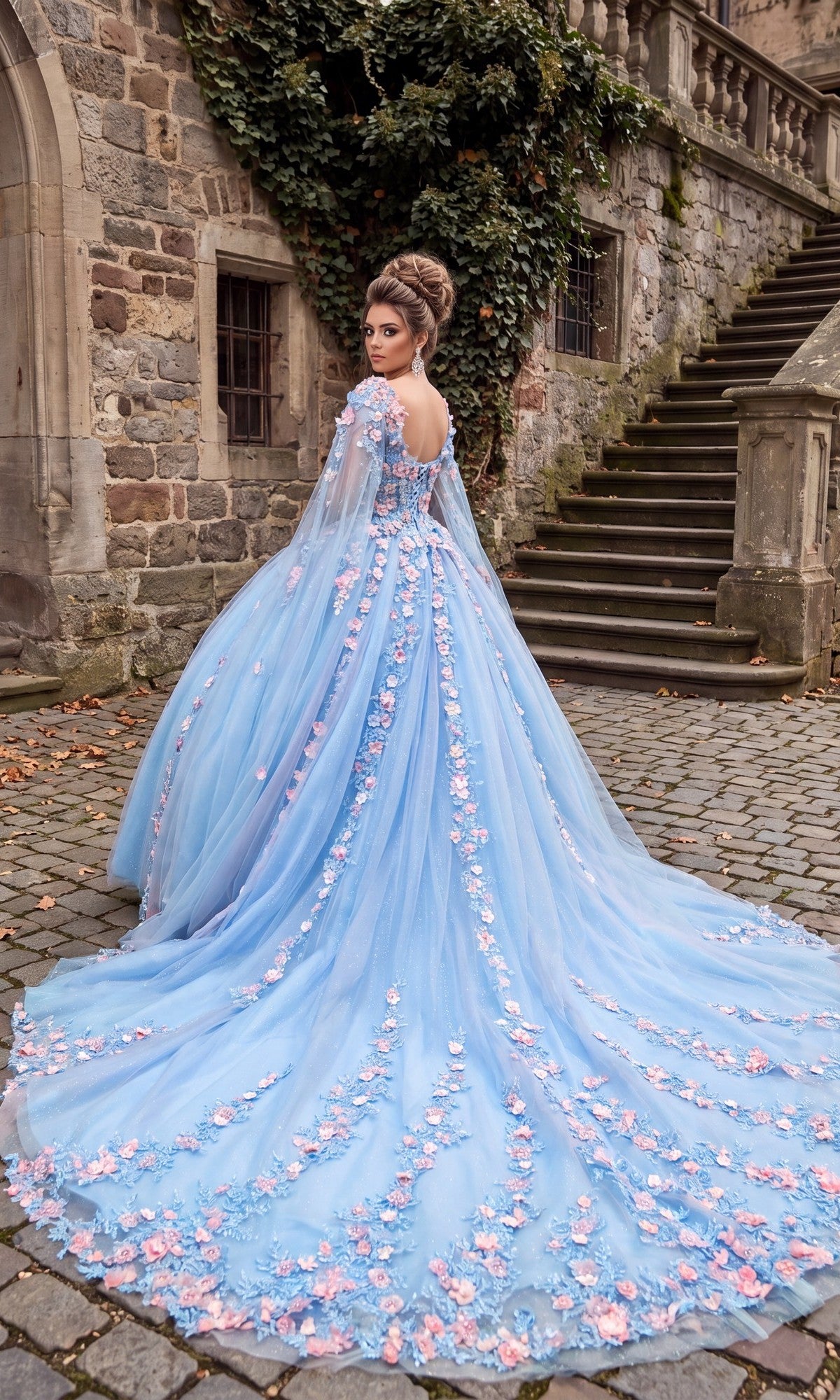 A woman wearing the Quinceanera Dress CHF3117 By Chicas—a long, light blue tulle ballgown adorned with pink 3D floral appliqués—stands on cobblestone near a stone building and staircase, looking over her shoulder.