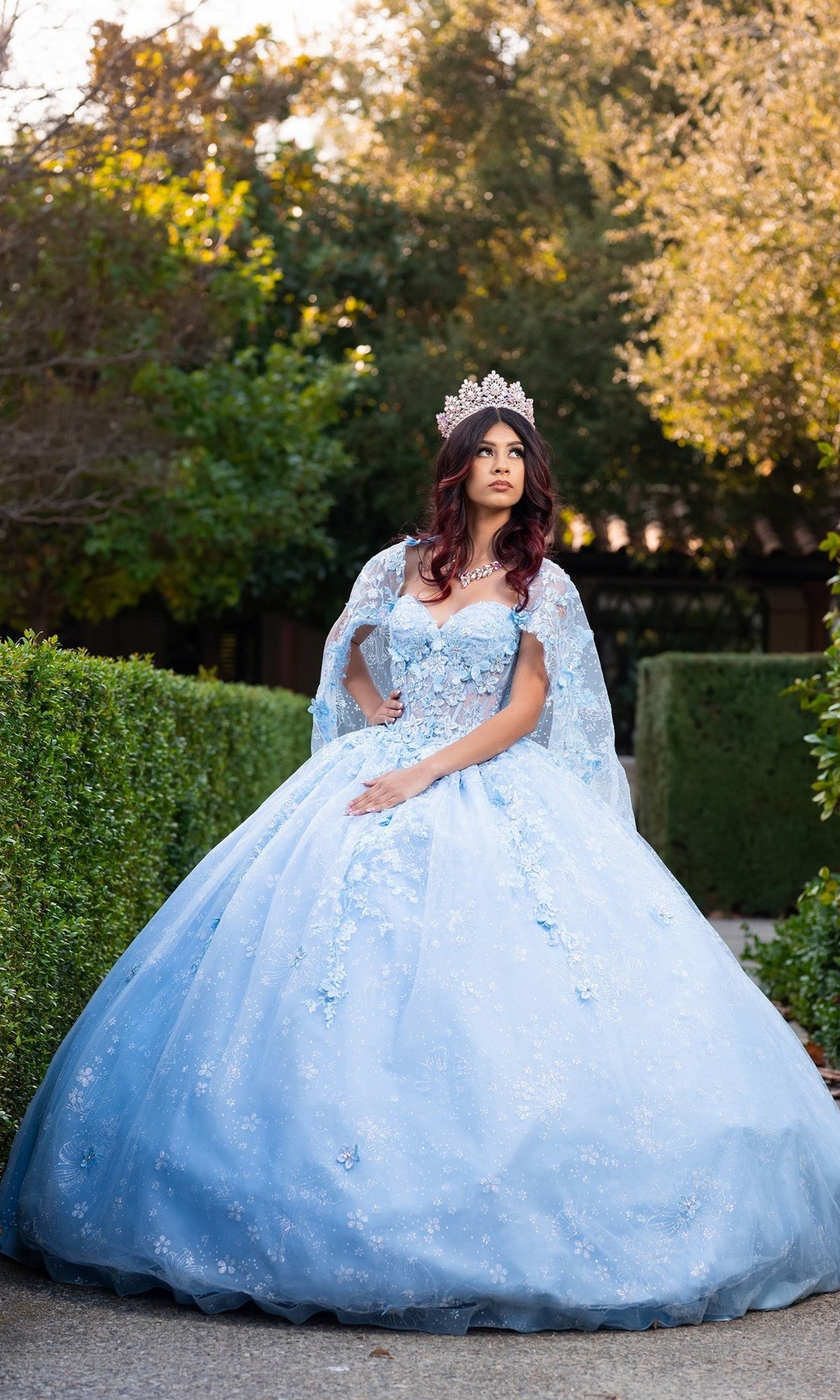 A woman stands outdoors on a garden path, surrounded by lush greenery and trees, wearing the Quinceanera Dress CHF3118 By Chicas in blue. The gown features delicate 3D floral appliques and a sparkling tiara, with its voluminous ballgown skirt adding an enchanting touch reminiscent of a pastel quinceañera look.