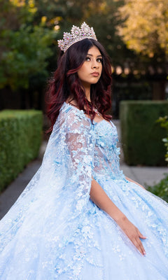 A woman stands outdoors among greenery and trees, looking off to the side, wearing the Quinceanera Dress CHF3118 By Chicas—a blue gown adorned with floral embroidery, 3D floral appliques, and a jeweled crown.