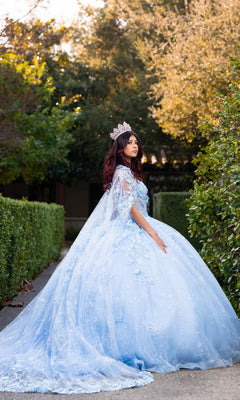 A woman stands on a path bordered by green hedges and trees, gazing to the side in the Quinceanera Dress CHF3118 By Chicas—a blue ball gown featuring a voluminous skirt and delicate 3D floral appliques.