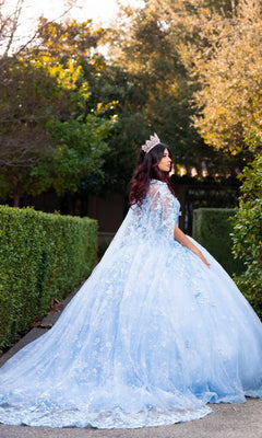 Wearing the Quinceanera Dress CHF3118 By Chicas—a light blue, elaborate gown with a voluminous ballgown skirt and delicate 3D floral appliques—a woman stands on a garden path surrounded by lush greenery and trees. Her jeweled crown enhances the regal charm of this enchanting scene.