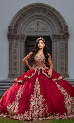 A young woman stands in front of an intricately carved stone doorway, wearing the Quinceanera Dress CHF3120 By Chicas—an ornate red and gold off-the-shoulder gown—along with a tiara as she looks ahead.