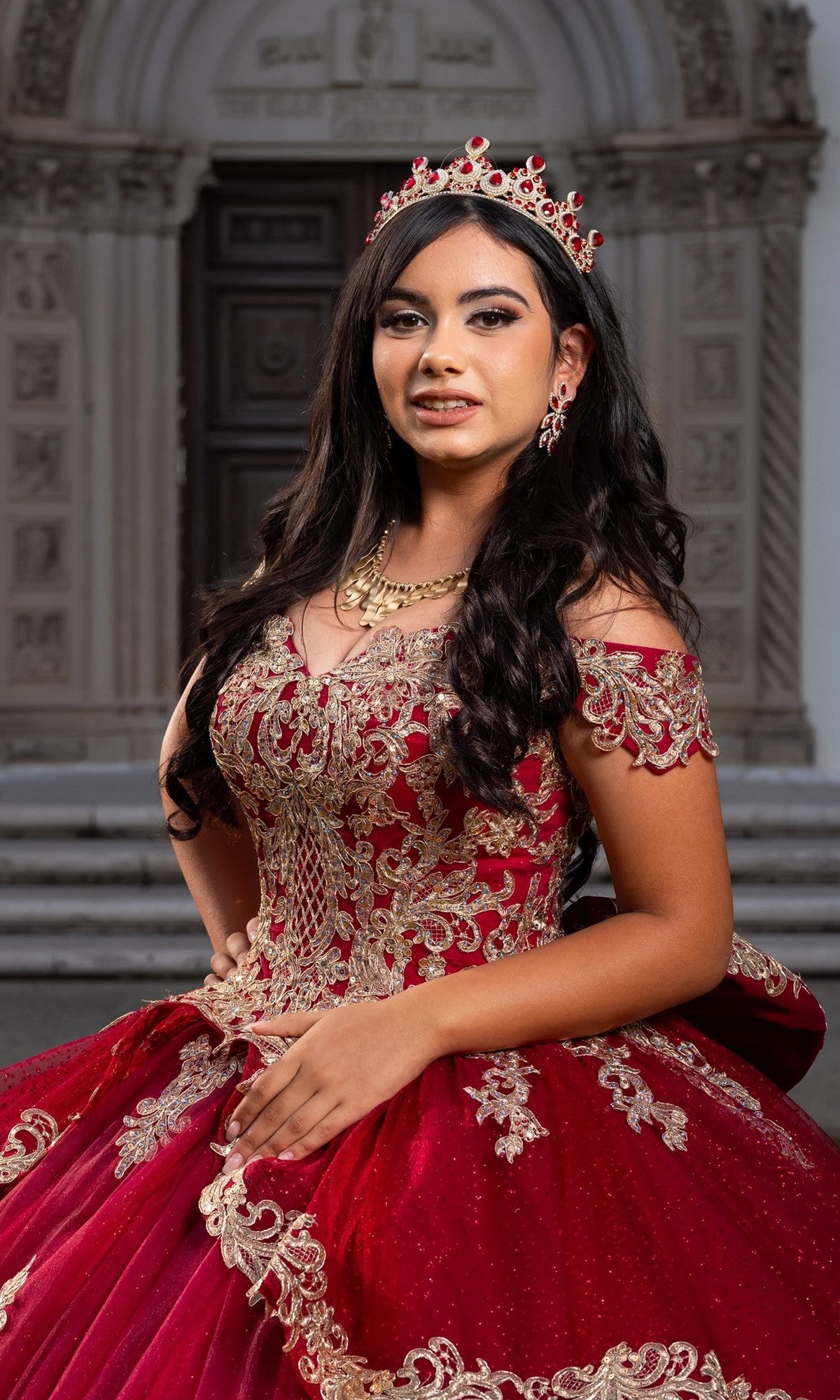 A young woman confidently poses at the entrance of a beautifully decorated building in the Quinceanera Dress CHF3120 By Chicas—an ornate red and gold off-the-shoulder gown adorned with exquisite gold embroidery.