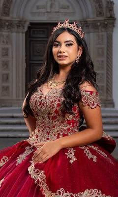 A young woman confidently poses at the entrance of a beautifully decorated building in the Quinceanera Dress CHF3120 By Chicas—an ornate red and gold off-the-shoulder gown adorned with exquisite gold embroidery.
