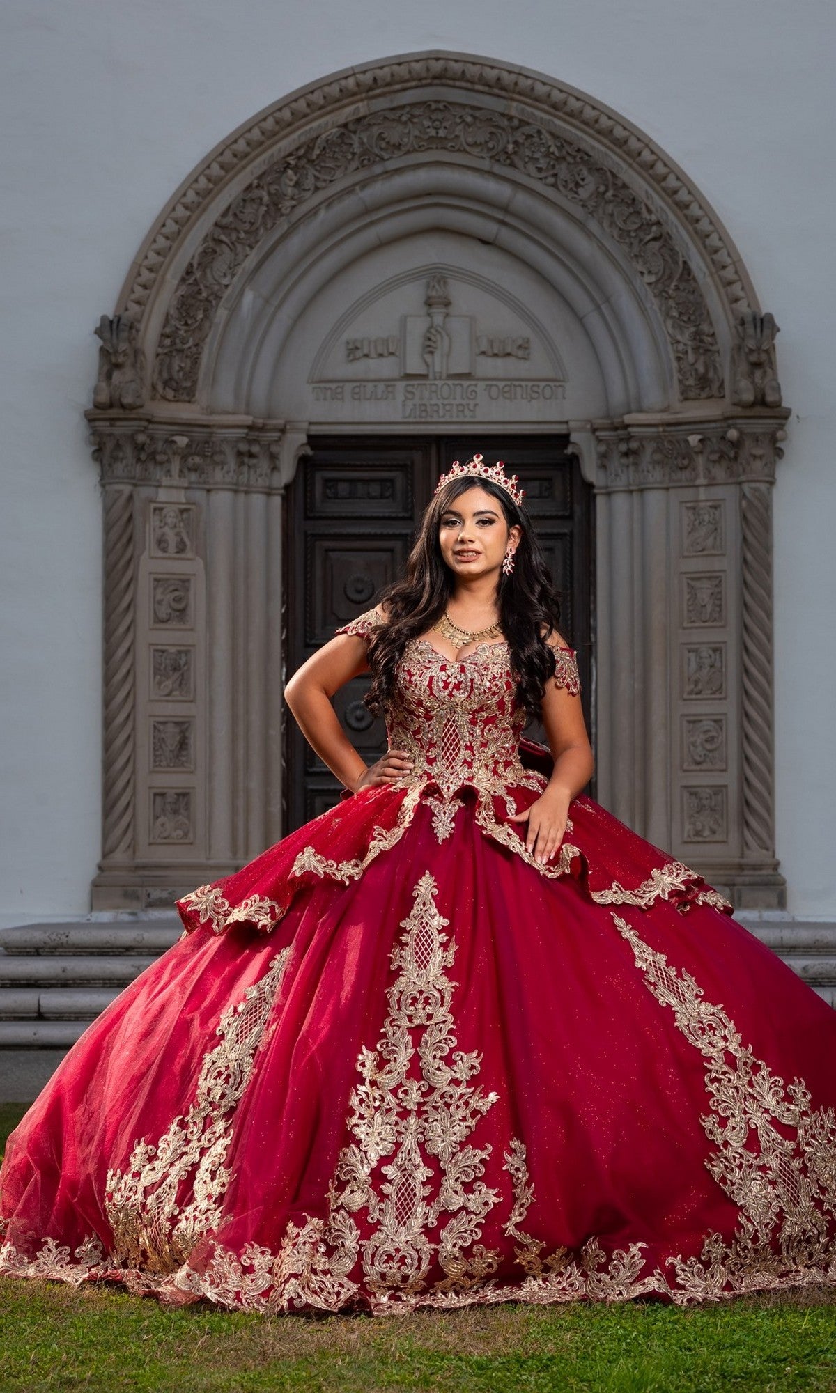 A young woman in the exquisite Quinceanera Dress CHF3120 By Chicas, featuring shimmering gold embroidery and glittering tulle in rich burgundy, stands regally before an ornate arched doorway, her crown gleaming above the embellished ball gown.