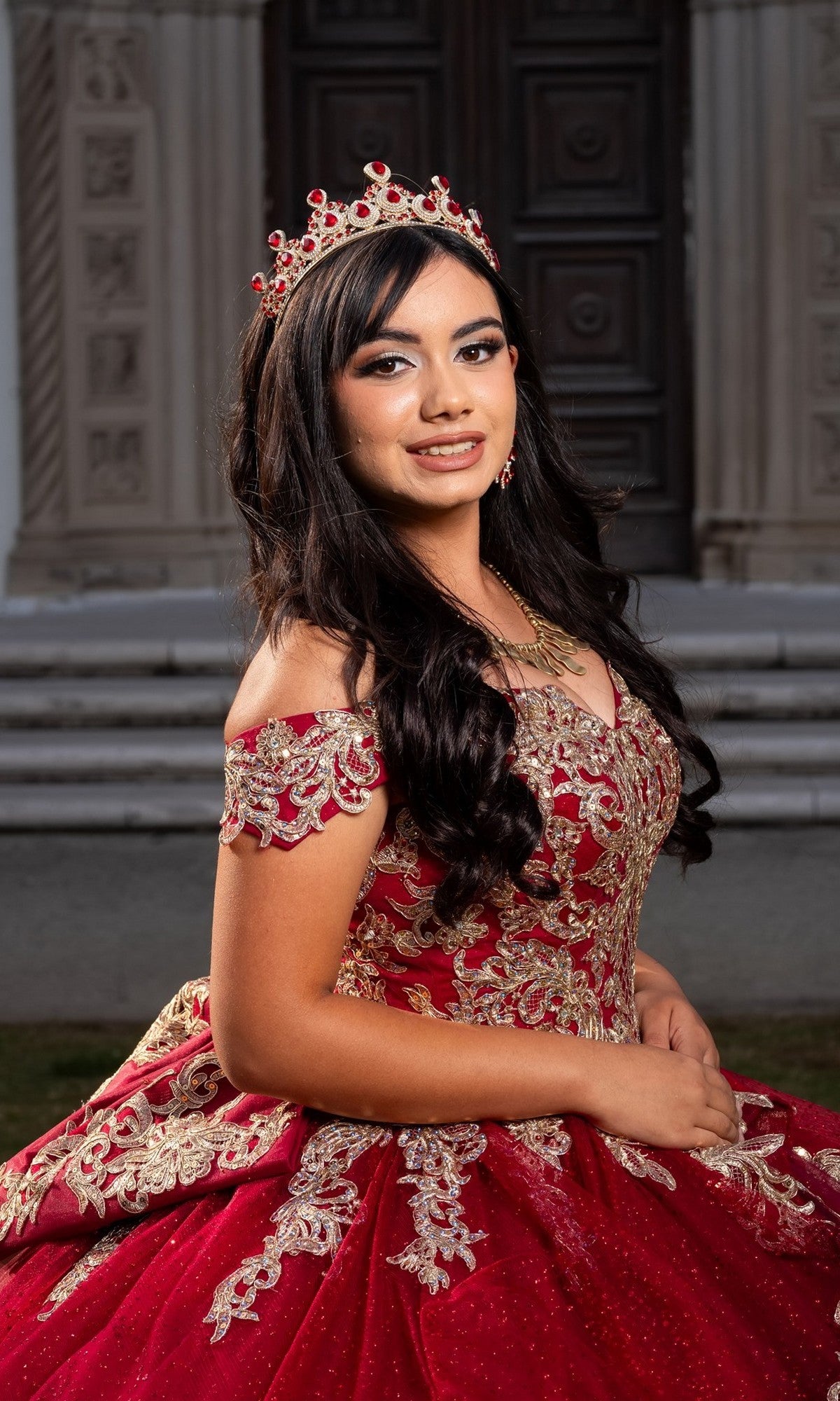 A young woman wearing the Quinceanera Dress CHF3120 By Chicas and a jeweled crown poses outdoors in front of a building with ornate doors.