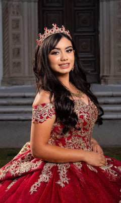 A young woman wearing the Quinceanera Dress CHF3120 By Chicas and a jeweled crown poses outdoors in front of a building with ornate doors.