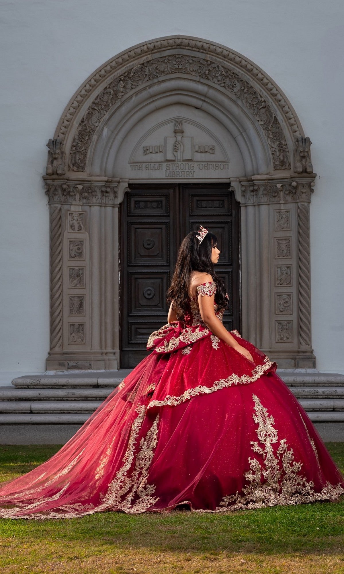 A young woman wearing the Quinceanera Dress CHF3120 By Chicas, an ornate off-the-shoulder gown featuring red and gold embroidery, stands sideways outside a large arched doorway with detailed stonework.