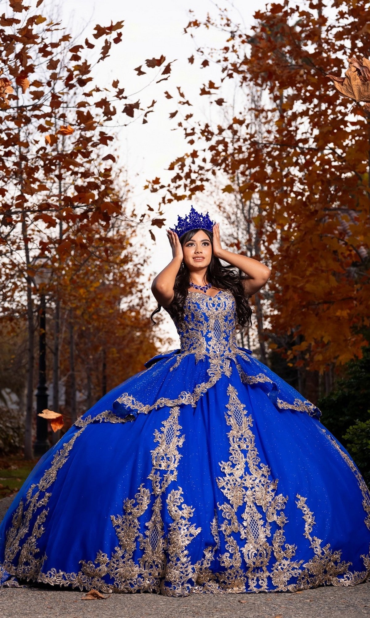 A young woman stands outdoors among autumn trees and scattered fallen leaves, wearing the Quinceanera Dress CHF3120 By Chicas—an ornate blue off-the-shoulder gown adorned with gold embroidery.