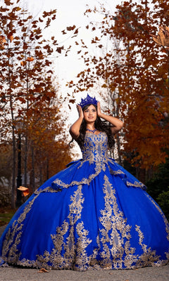 A young woman stands outdoors among autumn trees and scattered fallen leaves, wearing the Quinceanera Dress CHF3120 By Chicas—an ornate blue off-the-shoulder gown adorned with gold embroidery.
