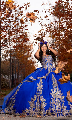 A young woman wearing the Quinceanera Dress CHF3120 By Chicas in royal poses outdoors on a path lined with autumn trees, smiling as large leaves fall around her.