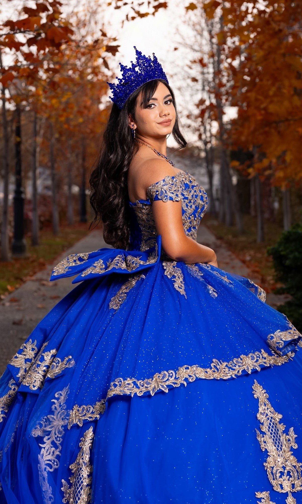 A young woman wearing the Quinceanera Dress CHF3120 By Chicas in royal blue and gold, paired with a matching crown, stands outdoors on an autumn path lined with trees.