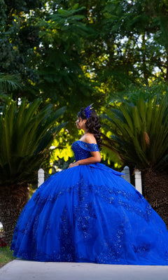 A young woman in a striking Quinceanera Dress CHF3122 By Chicas, featuring an ornate royal blue design, stands outdoors on a sidewalk surrounded by lush green plants and trees, facing to the right. The shimmering off-the-shoulder gown beautifully complements the vibrant scene.