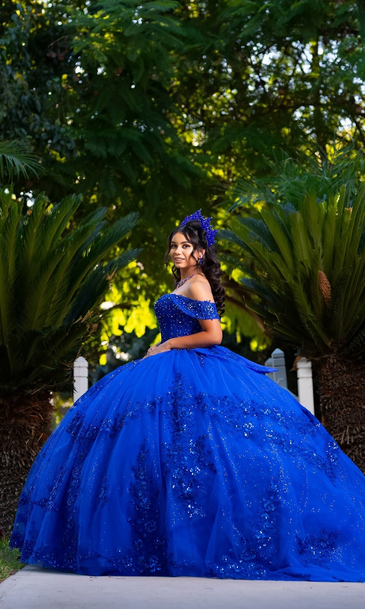 A young woman stands gracefully outdoors amid lush green plants, wearing the Quinceanera Dress CHF3122 By Chicas in royal blue. The elegant off-the-shoulder gown highlights her youthful beauty against the vibrant backdrop.