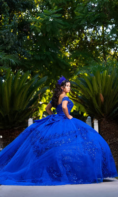 A young woman wearing the Quinceanera Dress CHF3122 by Chicas, featuring a royal blue, off-the-shoulder design and paired with a tiara, stands outdoors surrounded by vibrant green plants and trees.