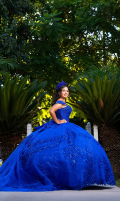 A young woman in the Quinceanera Dress CHF3122 by Chicas, a royal blue off-the-shoulder gown, poses outdoors against vibrant green foliage, gazing confidently at the camera.