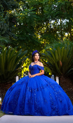 A woman stands outdoors among lush green plants, looking to the side in the stunning Quinceanera Dress CHF3122 By Chicas. The elegant off-the-shoulder design of this royal-blue gown highlights her graceful pose and creates a striking scene.