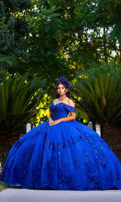 A young woman in the Quinceanera Dress CHF3122 by Chicas in royal poses outdoors between green palm bushes, wearing an elaborate off-the-shoulder gown and a matching headpiece.