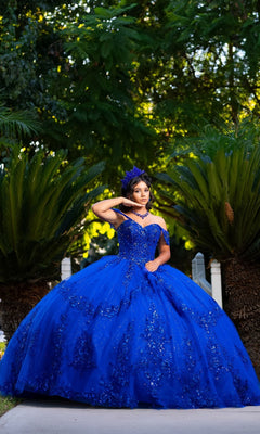 A young woman poses outdoors in the Quinceanera Dress CHF3122 By Chicas, a vibrant royal-blue ballgown adorned with floral embellishments. She completes her look with a matching headpiece and necklace. The off-the-shoulder design accentuates her elegant silhouette, set against a backdrop of lush green foliage.
