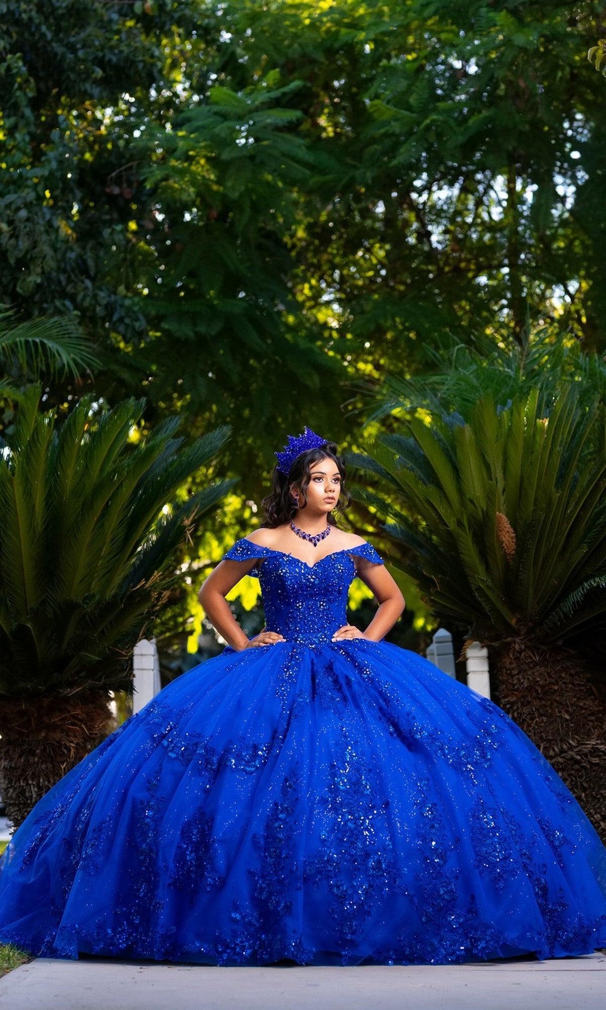 A young woman stands outdoors between two large green plants, hands on hips, wearing the Quinceanera Dress CHF3122 by Chicas—a stunning royal blue, off-the-shoulder ballgown adorned with beautiful embellishments—with lush greenery in the background.