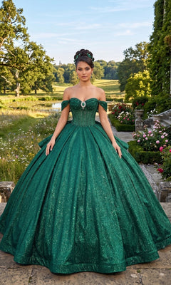 A woman wearing the Quinceanera Dress CHF3125 By Chicas in green, featuring an off-the-shoulder corset bodice, stands outdoors on stone steps with manicured gardens and trees in the background.