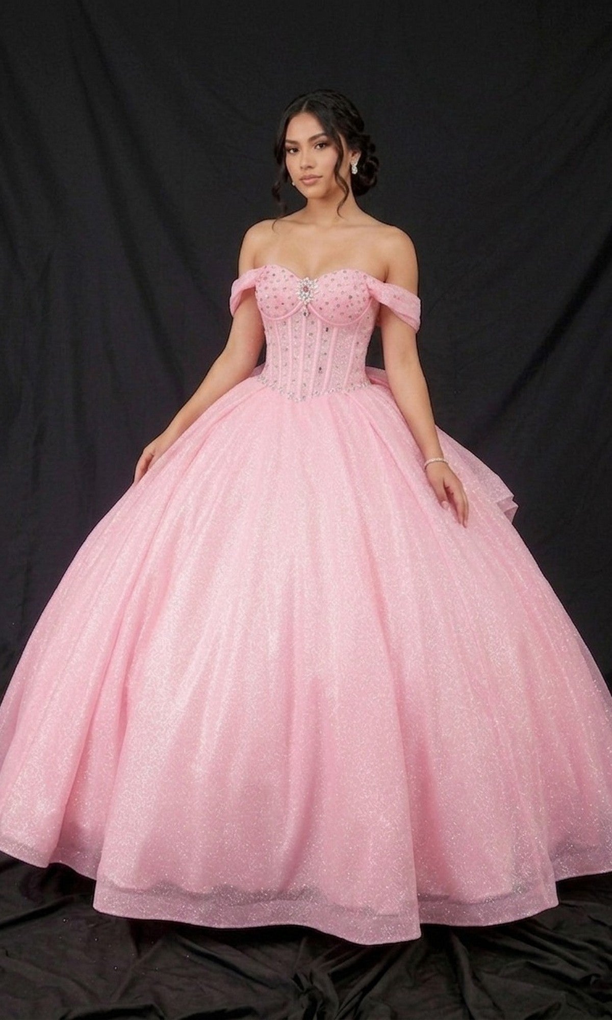 A young woman stands against a black backdrop wearing the Quinceanera Dress CHF3125 By Chicas, a pink glitter-tulle ballgown featuring an off-the-shoulder neckline, a fitted corset bodice, and a full skirt.