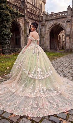 Wearing the Quinceanera Dress CHF3126 by Chicas, a woman stands on a cobblestone path outside a gothic-style stone building, looking over her shoulder. The off-the-shoulder ballgown features delicate floral embroidery that adds to its enchanting allure.