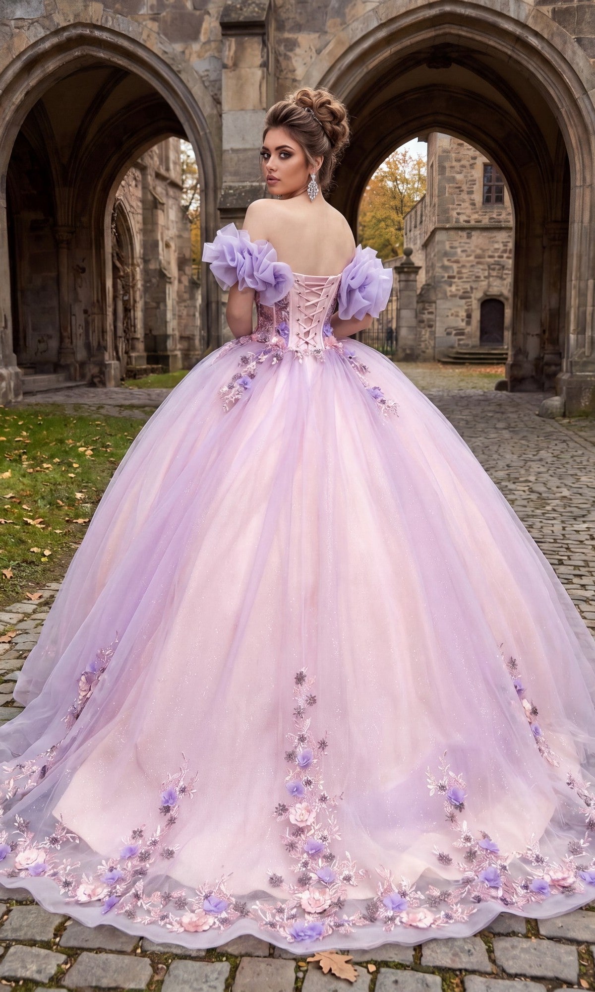 A woman stands on cobblestone in front of a stone building, her elegant ballgown skirt flowing behind as she faces away, wearing the Quinceanera Dress CHF3127 By Chicas—an off-the-shoulder lavender dress adorned with floral appliqués.
