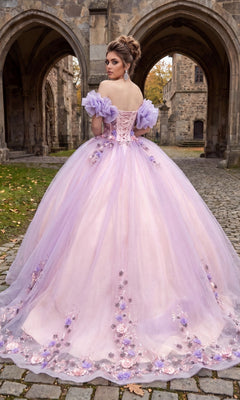 A woman stands on cobblestone in front of a stone building, her elegant ballgown skirt flowing behind as she faces away, wearing the Quinceanera Dress CHF3127 By Chicas—an off-the-shoulder lavender dress adorned with floral appliqués.