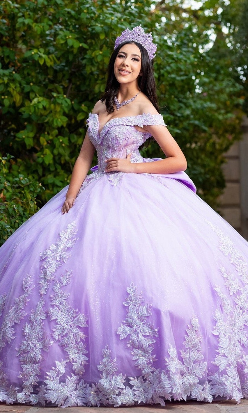 A young woman wearing the Quinceanera Dress CHF3128 By Chicas in lilac, adorned with delicate floral embroidery and a sparkling tiara, stands outdoors in front of green foliage, smiling at the camera.