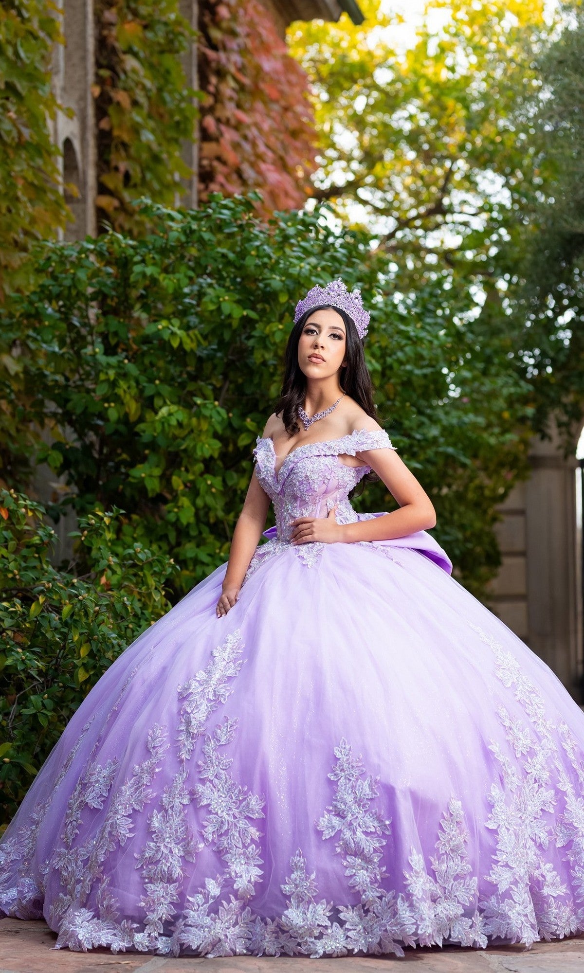 A young woman in the Quinceanera Dress CHF3128 by Chicas, featuring an off-the-shoulder design with delicate floral embroidery, stands outdoors in the sunlight surrounded by greenery, wearing a tiara and gazing off to the side.