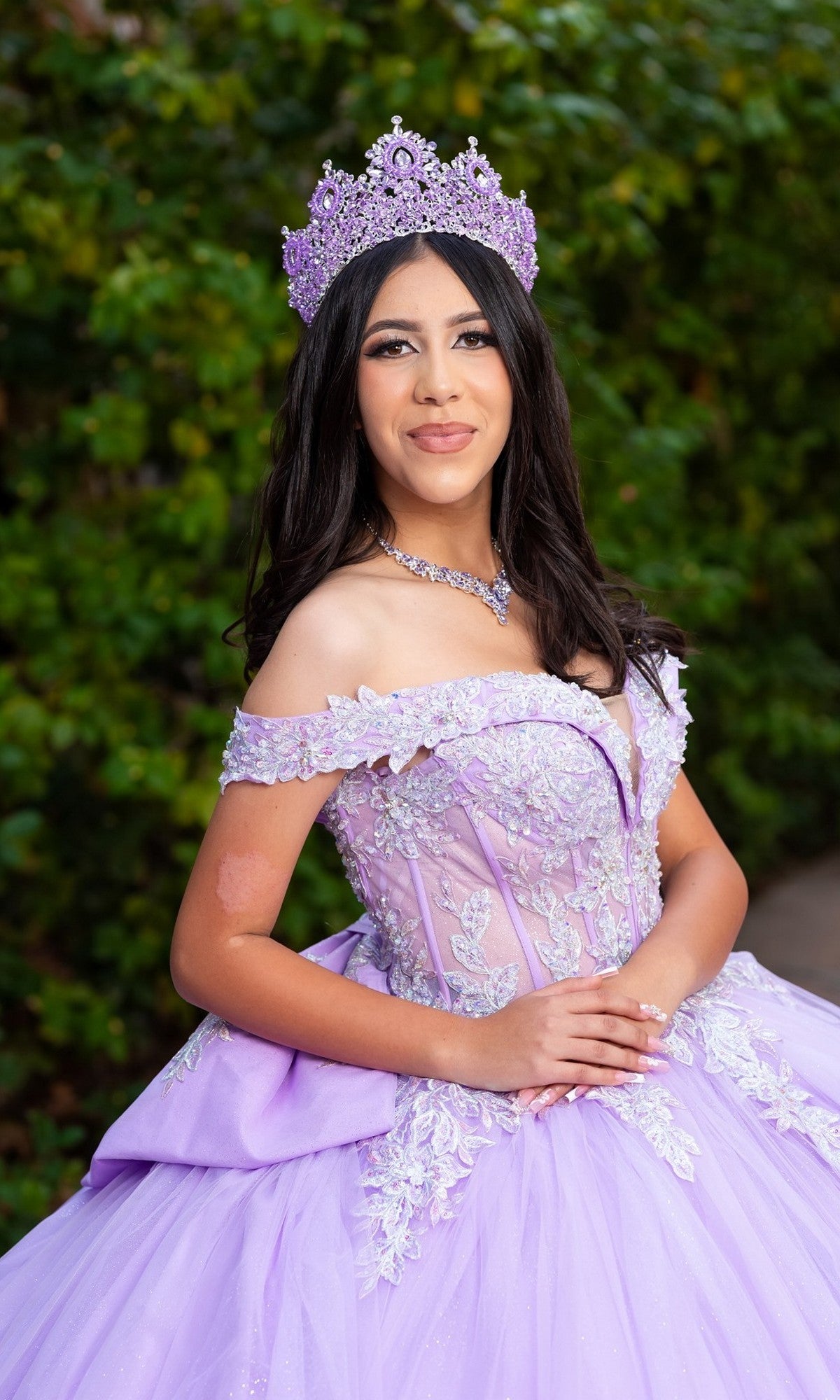 A young woman stands outdoors against lush green foliage, smiling with hands clasped, wearing the Quinceanera Dress CHF3128 By Chicas—a lilac gown adorned with delicate floral embroidery and a jeweled crown.