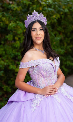A young woman stands outdoors against lush green foliage, smiling with hands clasped, wearing the Quinceanera Dress CHF3128 By Chicas—a lilac gown adorned with delicate floral embroidery and a jeweled crown.