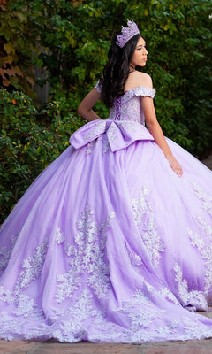 A young woman stands outdoors wearing the Quinceanera Dress CHF3128 By Chicas, a lilac gown adorned with elegant floral embroidery and a large bow, complemented by a matching crown.