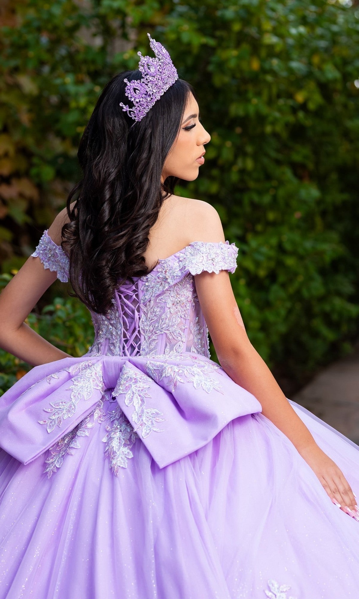 A young woman stands outdoors amid greenery, facing sideways in the Quinceanera Dress CHF3128 By Chicas. The lilac off-the-shoulder gown features intricate floral embroidery, a tiara, and a dramatic bow on the back.