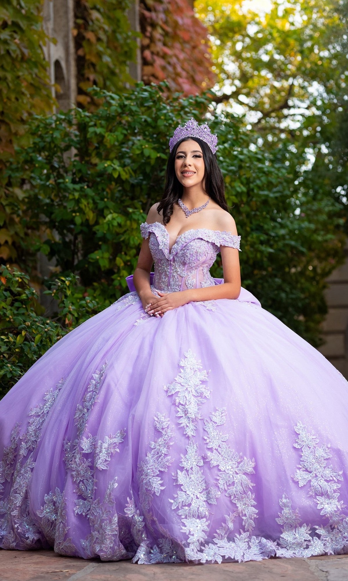 A young woman wearing the Quinceanera Dress CHF3128 By Chicas, which features an off-the-shoulder design with delicate floral embroidery, along with a crown and necklace, stands outdoors in front of greenery and a stone wall, smiling at the camera.