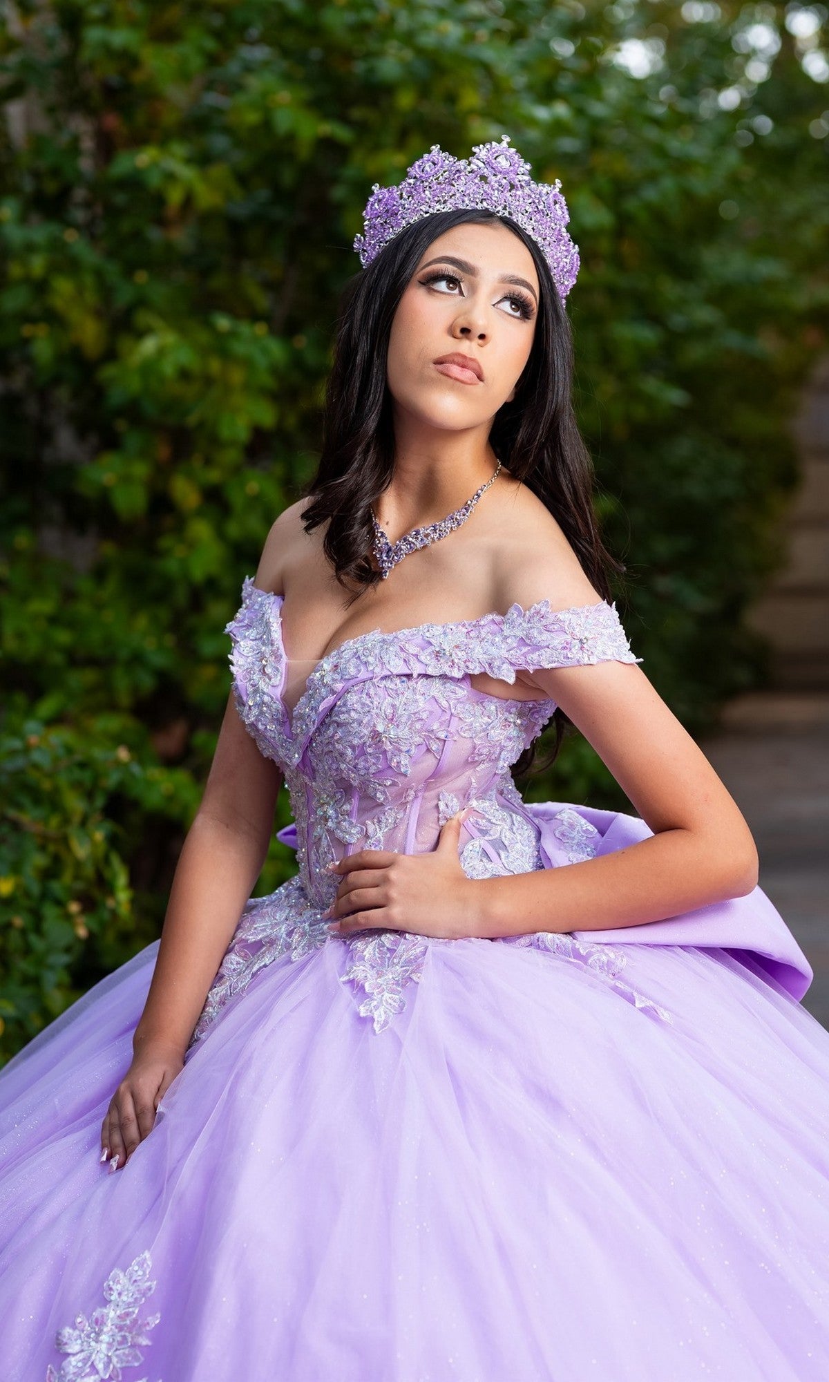 A young woman wearing the Quinceanera Dress CHF3128 By Chicas, an off-the-shoulder gown adorned with floral embroidery and paired with a matching tiara, poses outdoors against a backdrop of greenery.