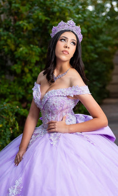 A young woman wearing the Quinceanera Dress CHF3128 By Chicas, an off-the-shoulder gown adorned with floral embroidery and paired with a matching tiara, poses outdoors against a backdrop of greenery.
