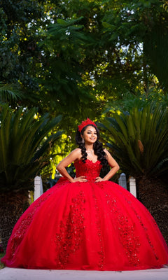 A young woman stands outdoors with her hands on her hips, wearing the Quinceanera Dress CHF3129 By Chicas—a bright red, embellished gown with sparkling glitter tulle and a matching crown—surrounded by green foliage.