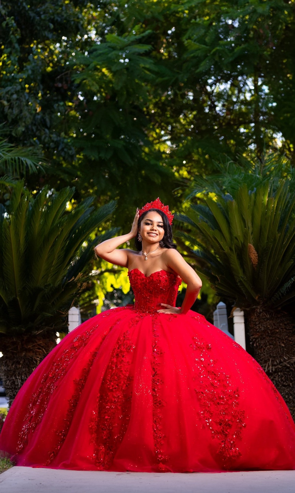 A young woman wearing the Quinceanera Dress CHF3129 By Chicas and a crown stands outdoors, smiling in front of green foliage.